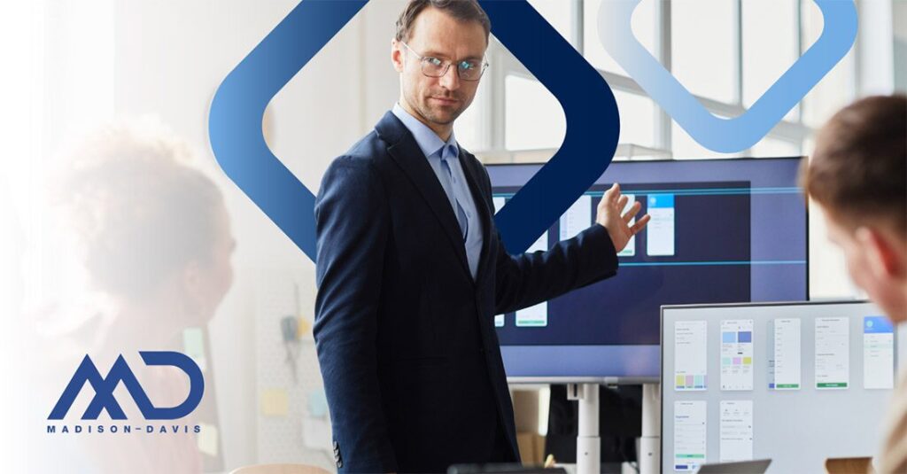 A man in a business suit and glasses speaks to two others, gesturing to a large computer monitor