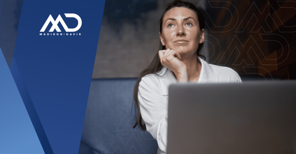 Viewed from behind a laptop screen, a woman in corporate attire pauses to think.