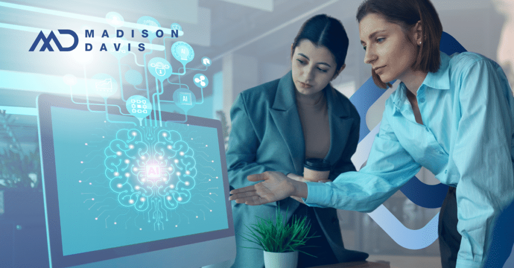 Two women in corporate attire view a computer monitor with a 2D digital brain labeled "AI"
